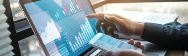 close up of a hand holding an iPhone and pointing at a screen filled with graphs