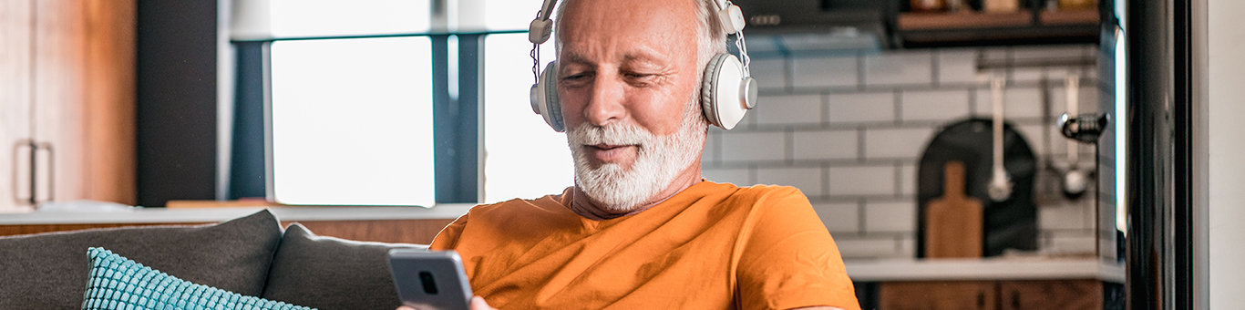 Older man sitting on a couch in his living room with headphones on, smiling at his phone